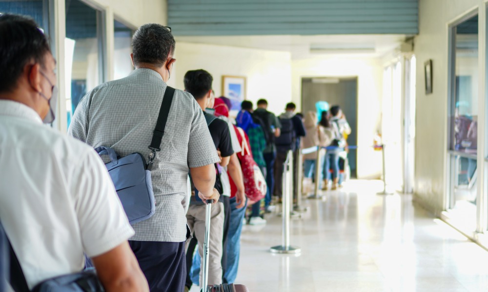 Travelers stand in a crowded airport terminal line with backpacks, luggage, seats, and digital signs nearby. Travelers stand in a crowded airport terminal line with backpacks, luggage, seats, and digital signs nearby.