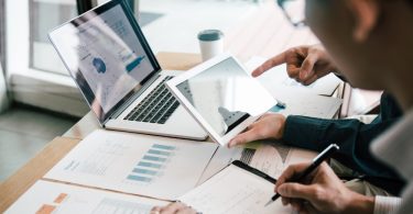 Business people in an office are reviewing charts on a laptop and tablet at a desk with printed reports next to them.