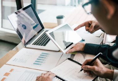 Business people in an office are reviewing charts on a laptop and tablet at a desk with printed reports next to them.