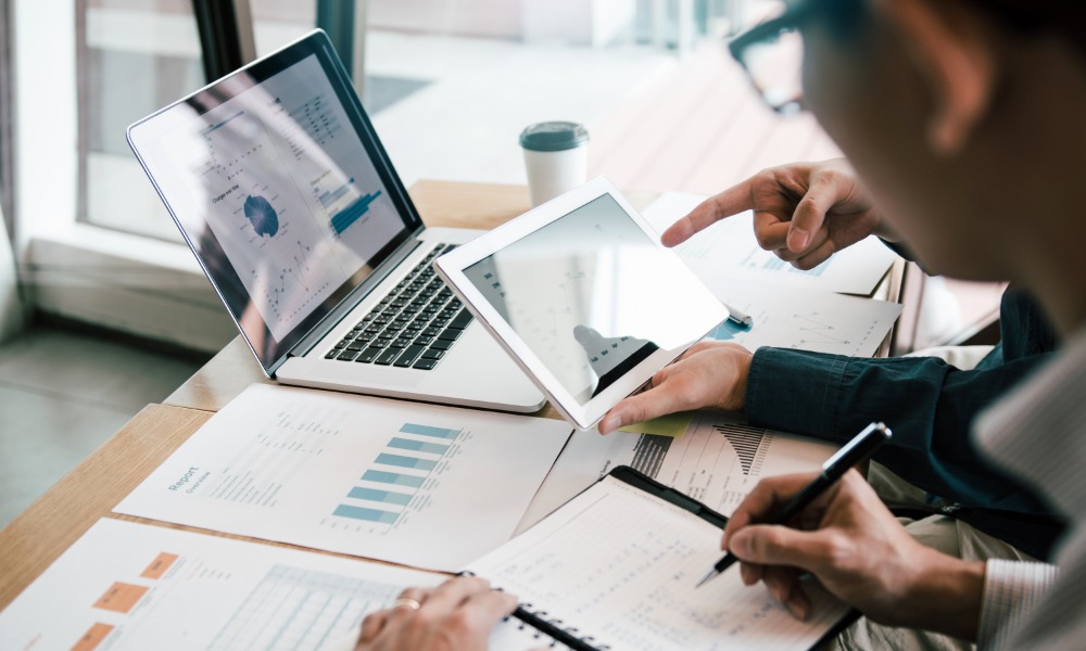 Business people in an office are reviewing charts on a laptop and tablet at a desk with printed reports next to them.