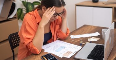 A woman in glasses sitting at a table with papers, a calculator phone, coins, bills, and an open laptop.