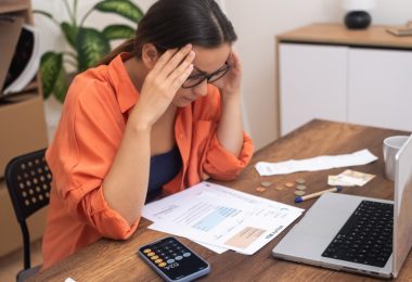 A woman in glasses sitting at a table with papers, a calculator phone, coins, bills, and an open laptop.