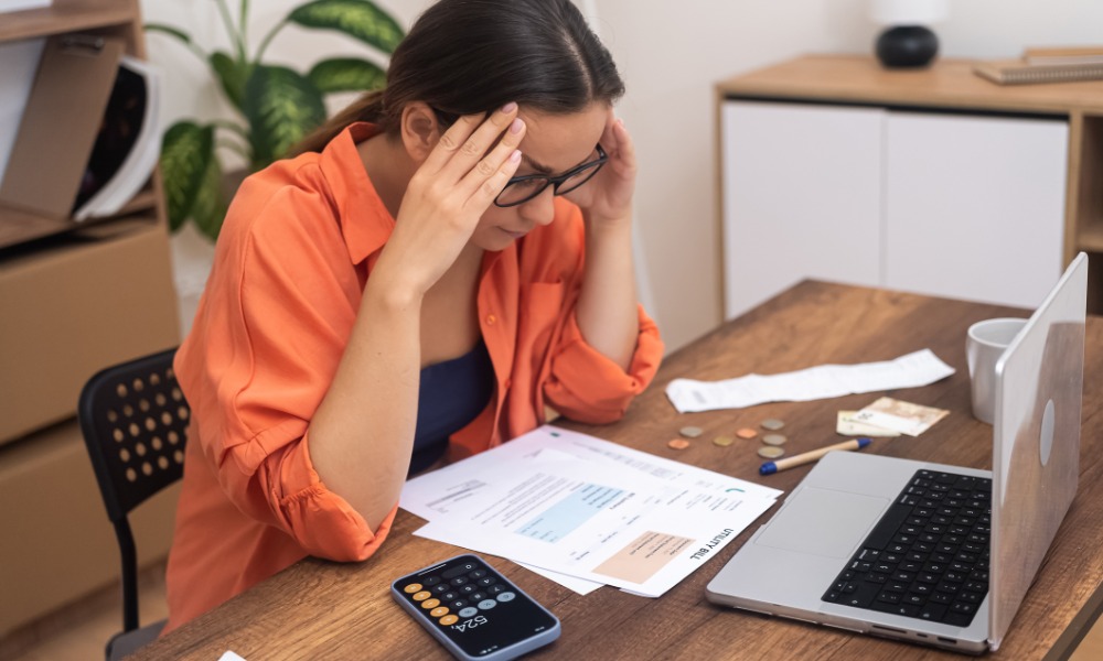 A woman in glasses sitting at a table with papers, a calculator phone, coins, bills, and an open laptop.