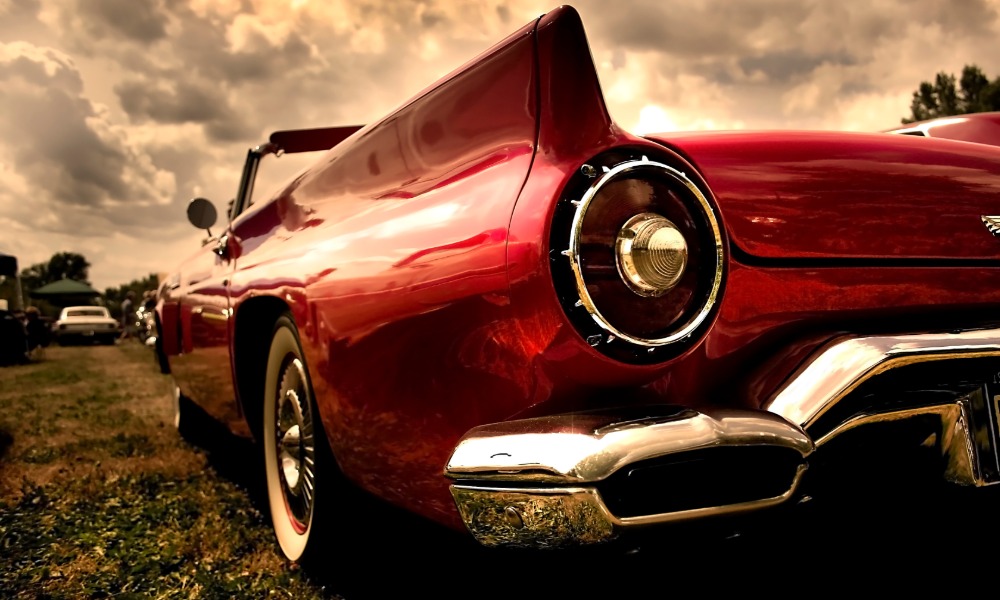 Low-angle view of a red classic convertible with chrome bumper parked outside beneath dramatic cloudy skies.