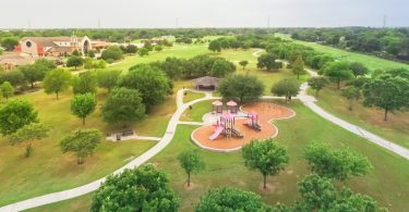 An aerial view of a community park with a playground, paved and clean sidewalks, and groomed grass and trees.
