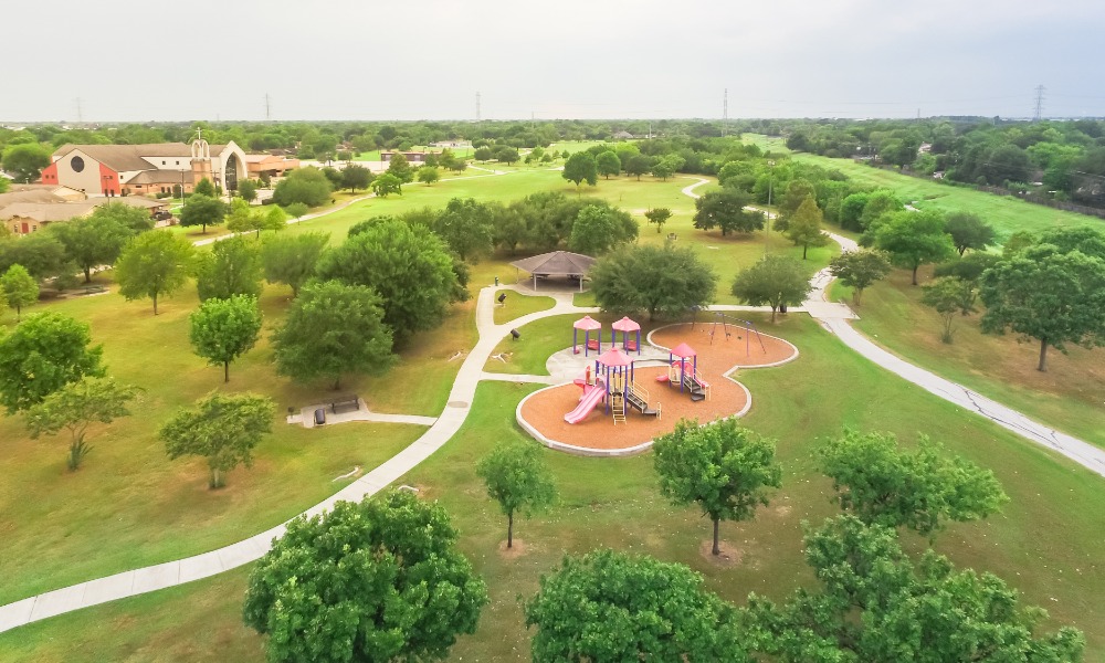 An aerial view of a community park with a playground, paved and clean sidewalks, and groomed grass and trees. An aerial view of a community park with a playground, paved and clean sidewalks, and groomed grass and trees.