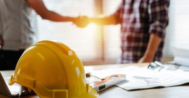 A close-up of a yellow hard hat resting on a desk. There are two people standing and shaking hands behind the desk.