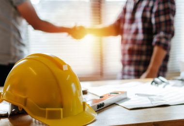 A close-up of a yellow hard hat resting on a desk. There are two people standing and shaking hands behind the desk.