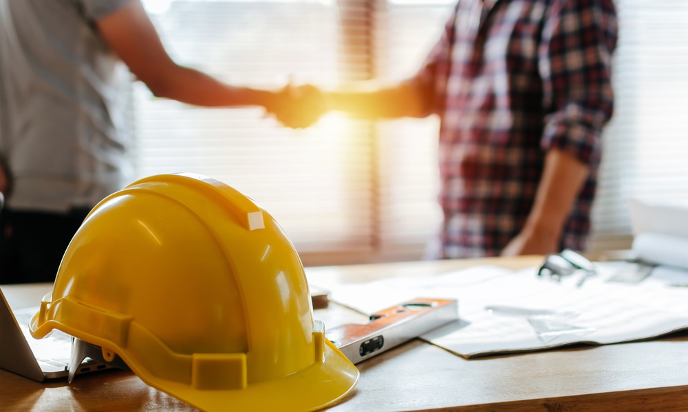 A close-up of a yellow hard hat resting on a desk. There are two people standing and shaking hands behind the desk.