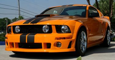 An orange sports car with black racing stripes, chrome wheels, a hood scoop, and a prominent front grille.