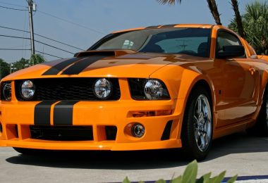 An orange sports car with black racing stripes, chrome wheels, a hood scoop, and a prominent front grille.
