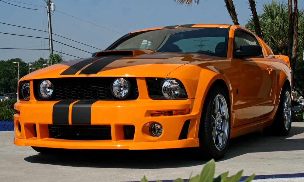 An orange sports car with black racing stripes, chrome wheels, a hood scoop, and a prominent front grille.