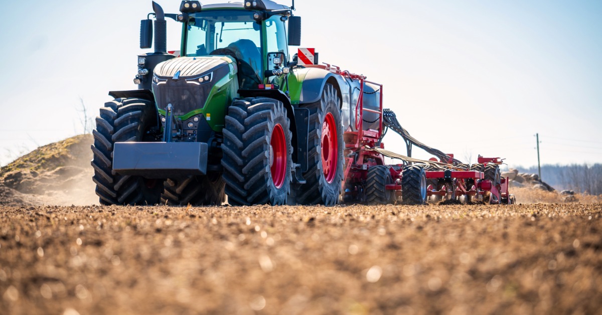 A green tractor with large tires pulling a red seeder across a tilled field with loose soil and dust behind it.