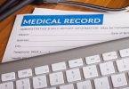 A stethoscope and pen resting on health insurance paperwork and a medical record folder on a desk with white paper beneath.
