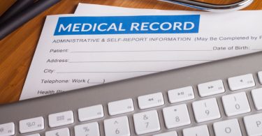 A stethoscope and pen resting on health insurance paperwork and a medical record folder on a desk with white paper beneath.