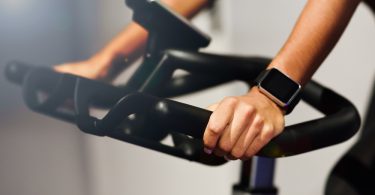 A person gripping indoor cycling bike handlebars while wearing a dark smartwatch during exercise indoors.