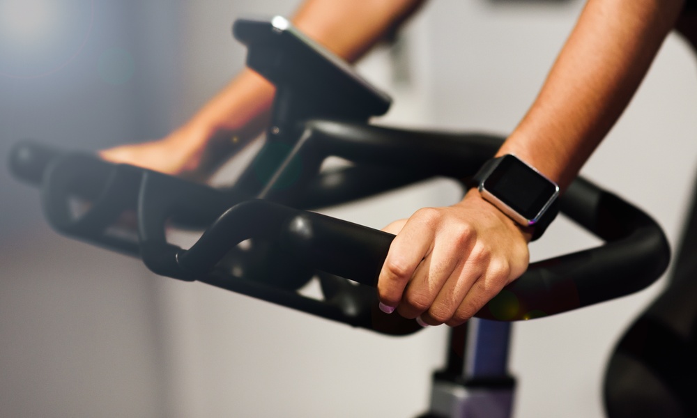 A person gripping indoor cycling bike handlebars while wearing a dark smartwatch during exercise indoors.
