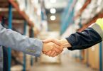 A businessperson and a warehouse worker in a high-visibility jacket shaking hands between warehouse shelves.
