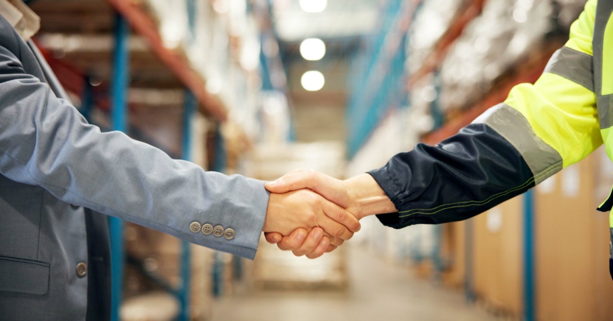 A businessperson and a warehouse worker in a high-visibility jacket shaking hands between warehouse shelves.