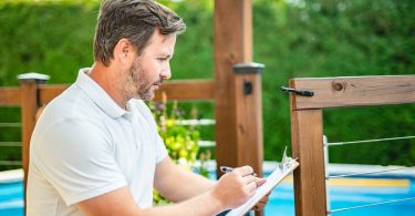 A person writing on a clipboard beside a wooden pool safety fence with blue water and greenery behind it.