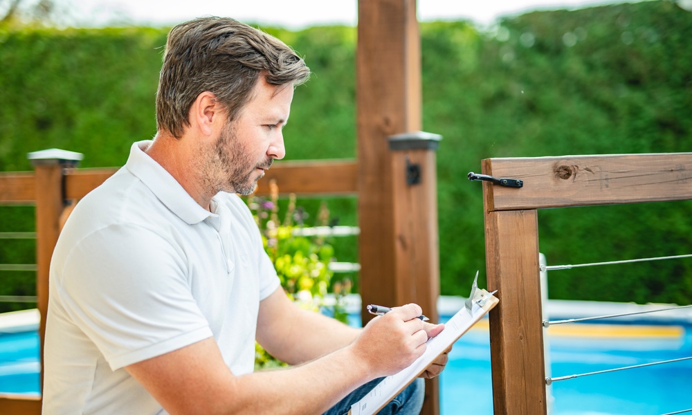 A person writing on a clipboard beside a wooden pool safety fence with blue water and greenery behind it.