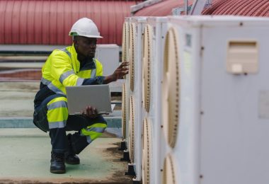 A man is kneeling next to several air conditioning units. The man has a laptop in one hand and is wearing a hard hat.