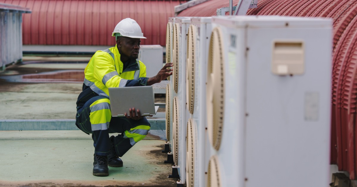 A man is kneeling next to several air conditioning units. The man has a laptop in one hand and is wearing a hard hat.