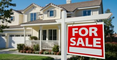A two-story house with trees and a grassy lawn features a sign on its front lawn that reads "For Sale" in big letters.