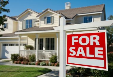 A two-story house with trees and a grassy lawn features a sign on its front lawn that reads "For Sale" in big letters.