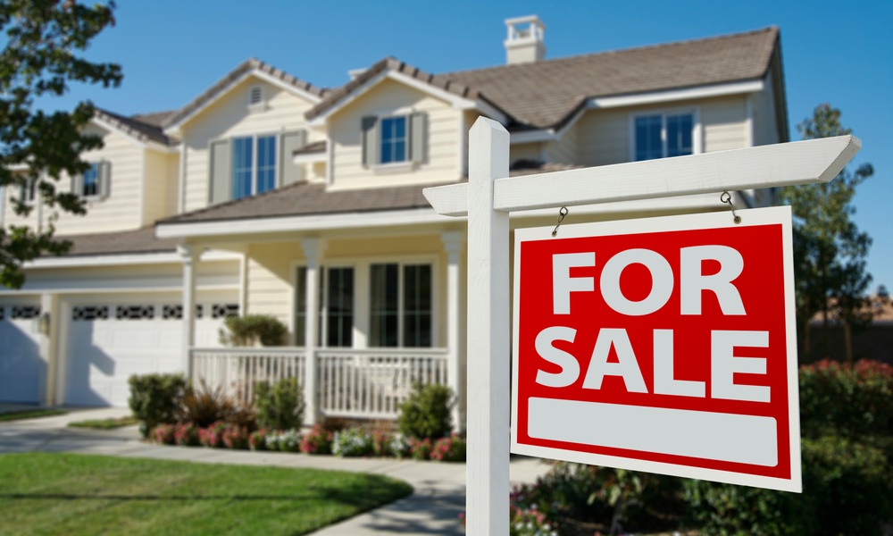 A two-story house with trees and a grassy lawn features a sign on its front lawn that reads "For Sale" in big letters.