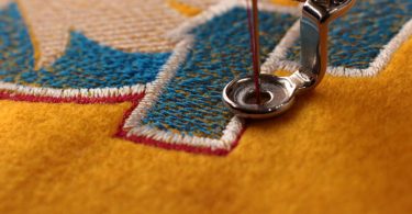 A sewing needle moves through a presser foot. It stitches red thread around a blue-and-white embroidery design.