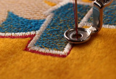A sewing needle moves through a presser foot. It stitches red thread around a blue-and-white embroidery design.