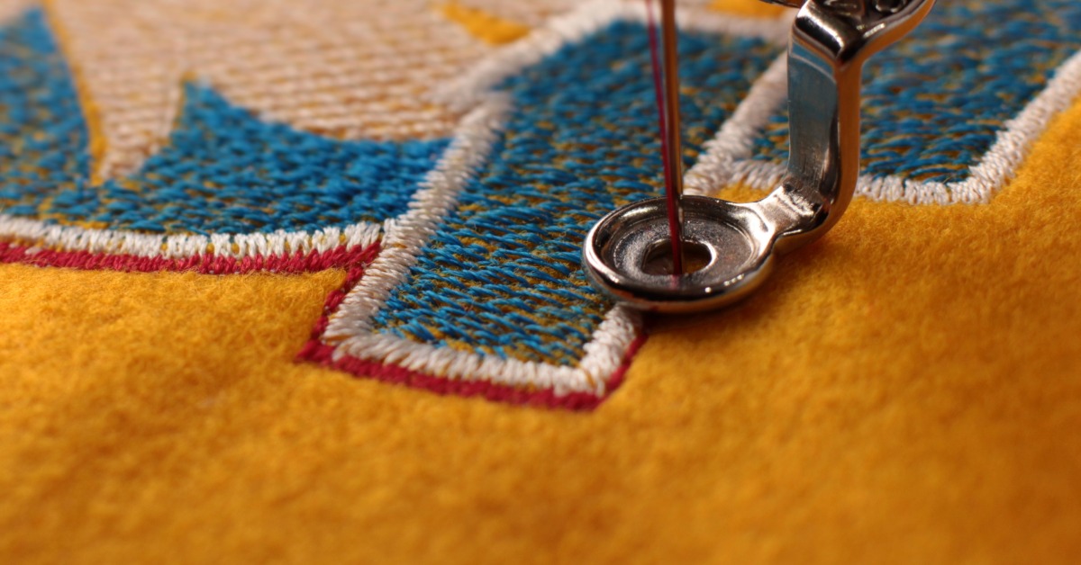 A sewing needle moves through a presser foot. It stitches red thread around a blue-and-white embroidery design.