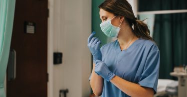 Healthcare worker puts on a blue glove while wearing scrubs and a face mask in a clinical setting.