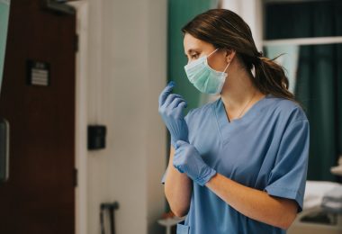Healthcare worker puts on a blue glove while wearing scrubs and a face mask in a clinical setting.