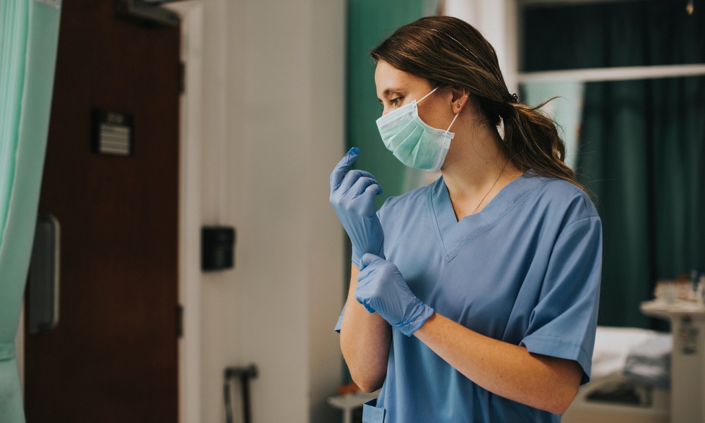 Healthcare worker puts on a blue glove while wearing scrubs and a face mask in a clinical setting.