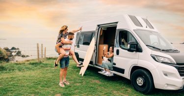 A woman in a camper van taking a photo of one person standing with a girl on his back with the coastline behind them.