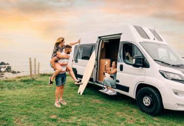 A woman in a camper van taking a photo of one person standing with a girl on his back with the coastline behind them.
