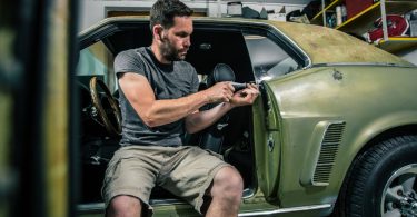 A man in the driver's seat of an classic, green, sports car with the door open and fixing the trim panels with a tool.