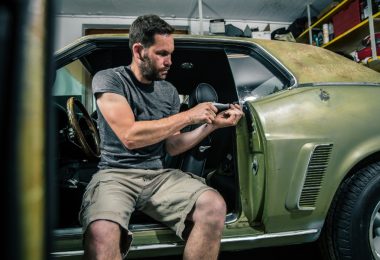 A man in the driver's seat of an classic, green, sports car with the door open and fixing the trim panels with a tool.