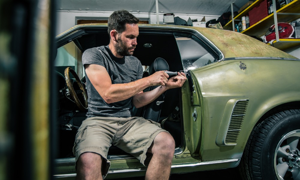 A man in the driver's seat of an classic, green, sports car with the door open and fixing the trim panels with a tool.