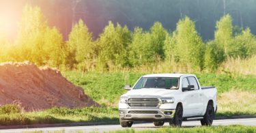 A white pickup truck with four doors and black wheels, driving on a concrete road between greenery and a dirt pile.
