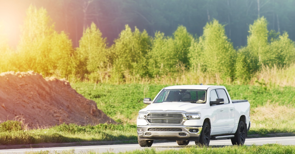 A white pickup truck with four doors and black wheels, driving on a concrete road between greenery and a dirt pile.