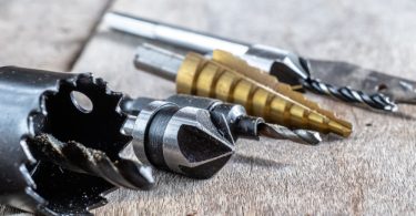 Close-up of drill bits, a hole saw, and cutting attachments laid out on a weathered wooden work surface.