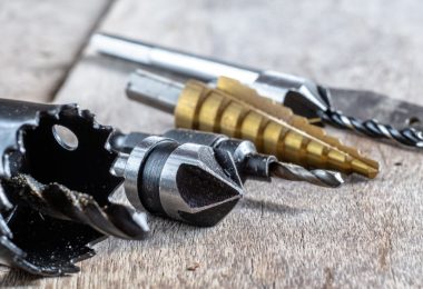Close-up of drill bits, a hole saw, and cutting attachments laid out on a weathered wooden work surface.