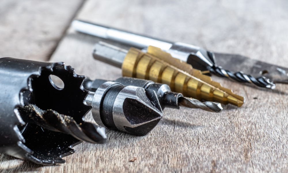 Close-up of drill bits, a hole saw, and cutting attachments laid out on a weathered wooden work surface.