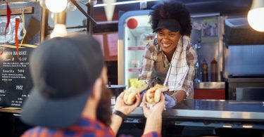 A young woman leaning out the window of a food truck handing out hot dogs to a customer right outside.