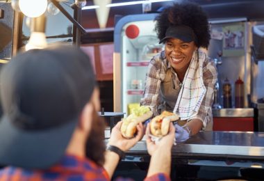 A young woman leaning out the window of a food truck handing out hot dogs to a customer right outside.