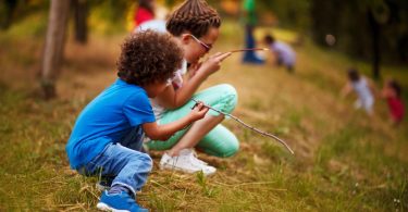Two children are in a forested area, kneeling on the ground and looking at sticks. There are more kids in the background.