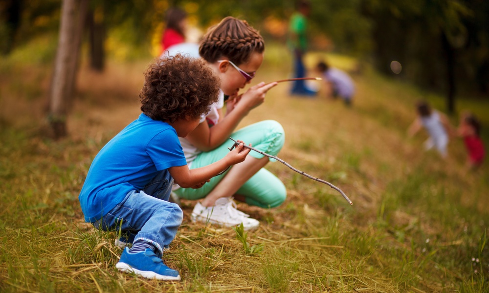 Two children are in a forested area, kneeling on the ground and looking at sticks. There are more kids in the background.